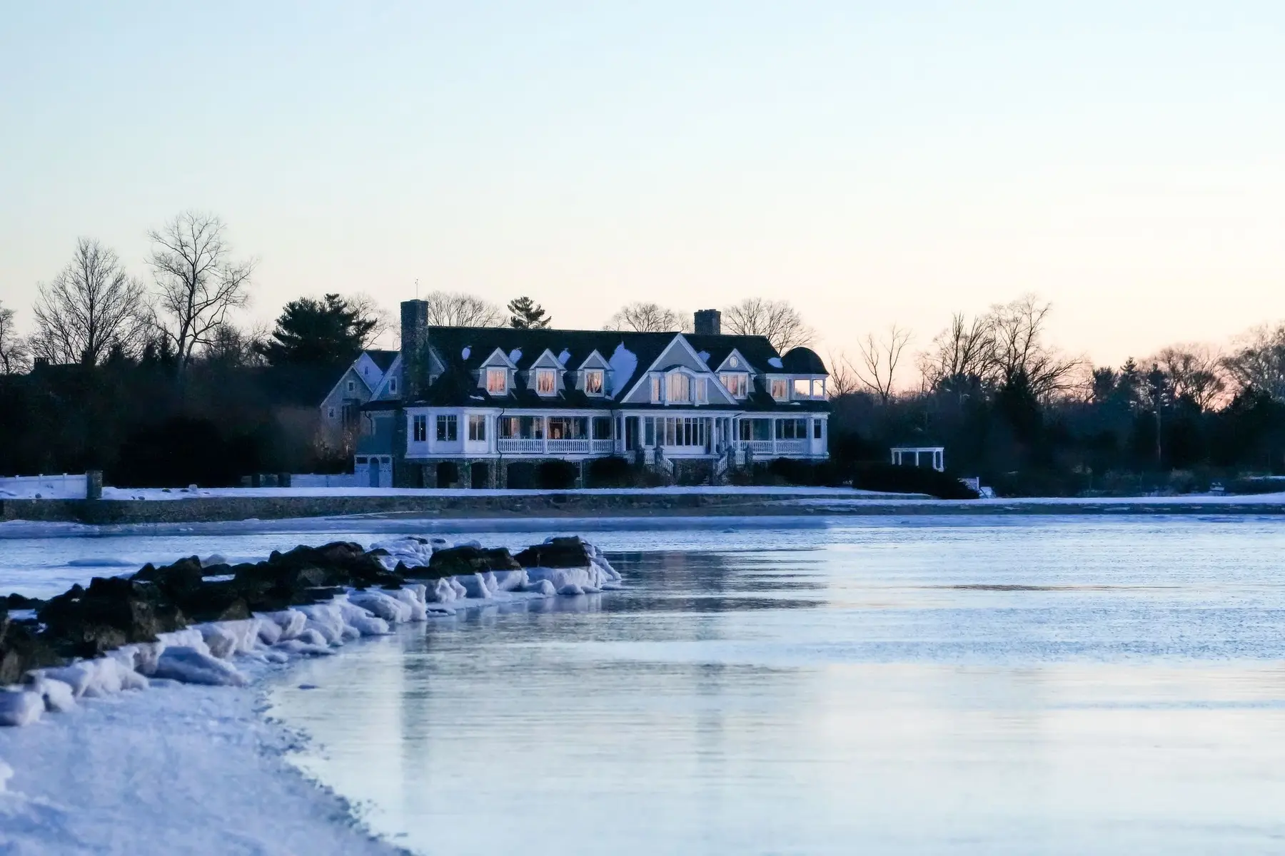 A shingle-style waterfront estate under snow at dusk, its windows lit — a Hamptons second home watched over through the off-season.