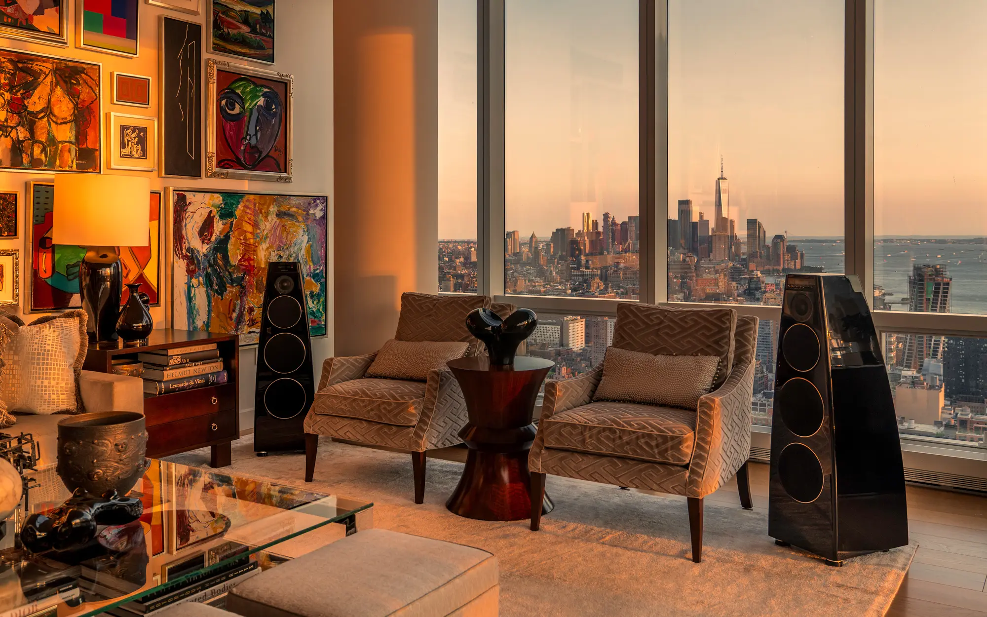 A penthouse living room at golden hour, floorstanding loudspeakers beside floor-to-ceiling glass over the city