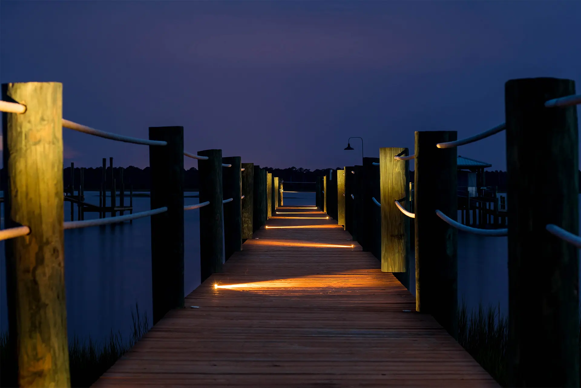 A lit dock reaching over the water at dusk, lined with warm Coastal Source path lighting